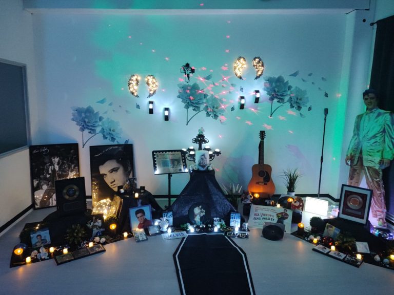 Ein dekorierter Raum mit Fotos, Kerzen und einer Gitarre, umgeben von buntem Licht.