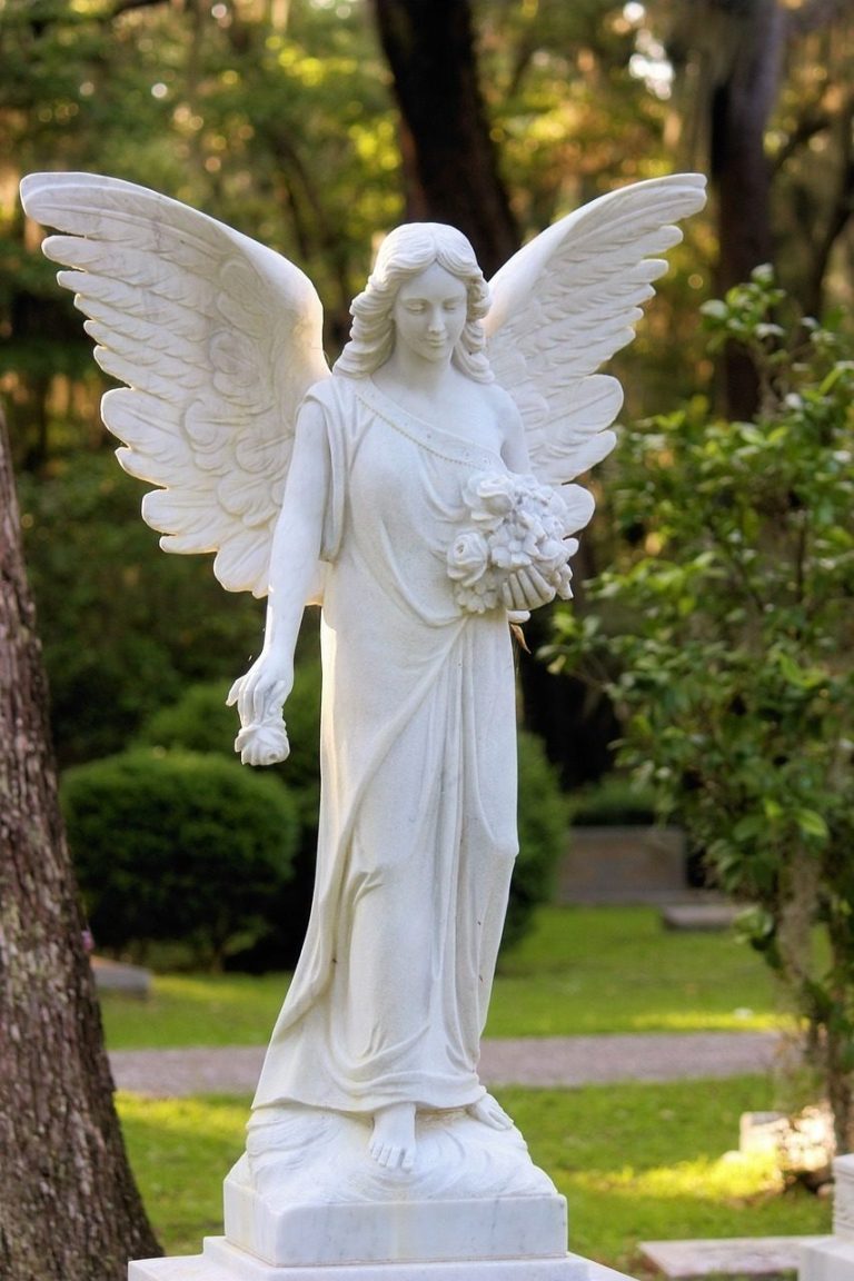 Eine weiße Engelsstatue mit Flügeln, die Blumen hält, umgeben von Bäumen im Freien.